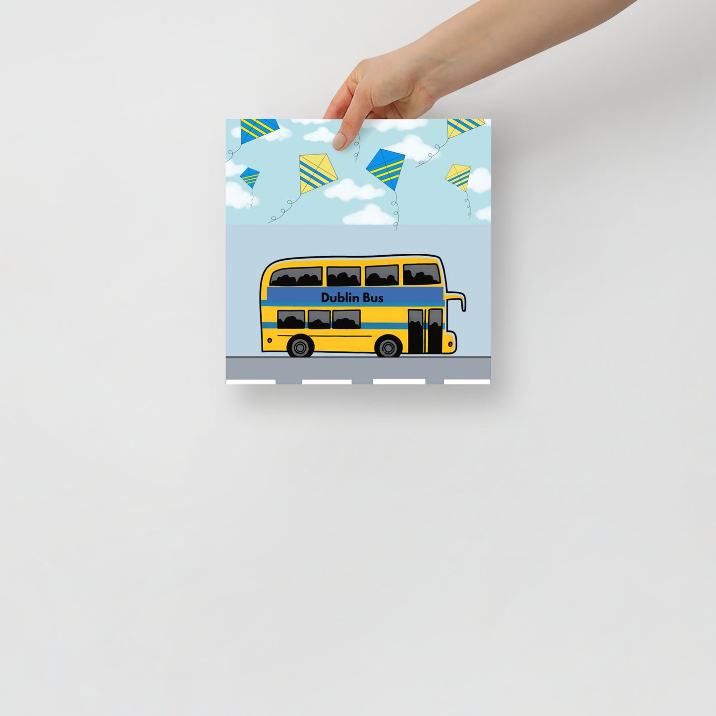 Hand holding an art poster with a yellow dublin bus design and kites on a light grey background