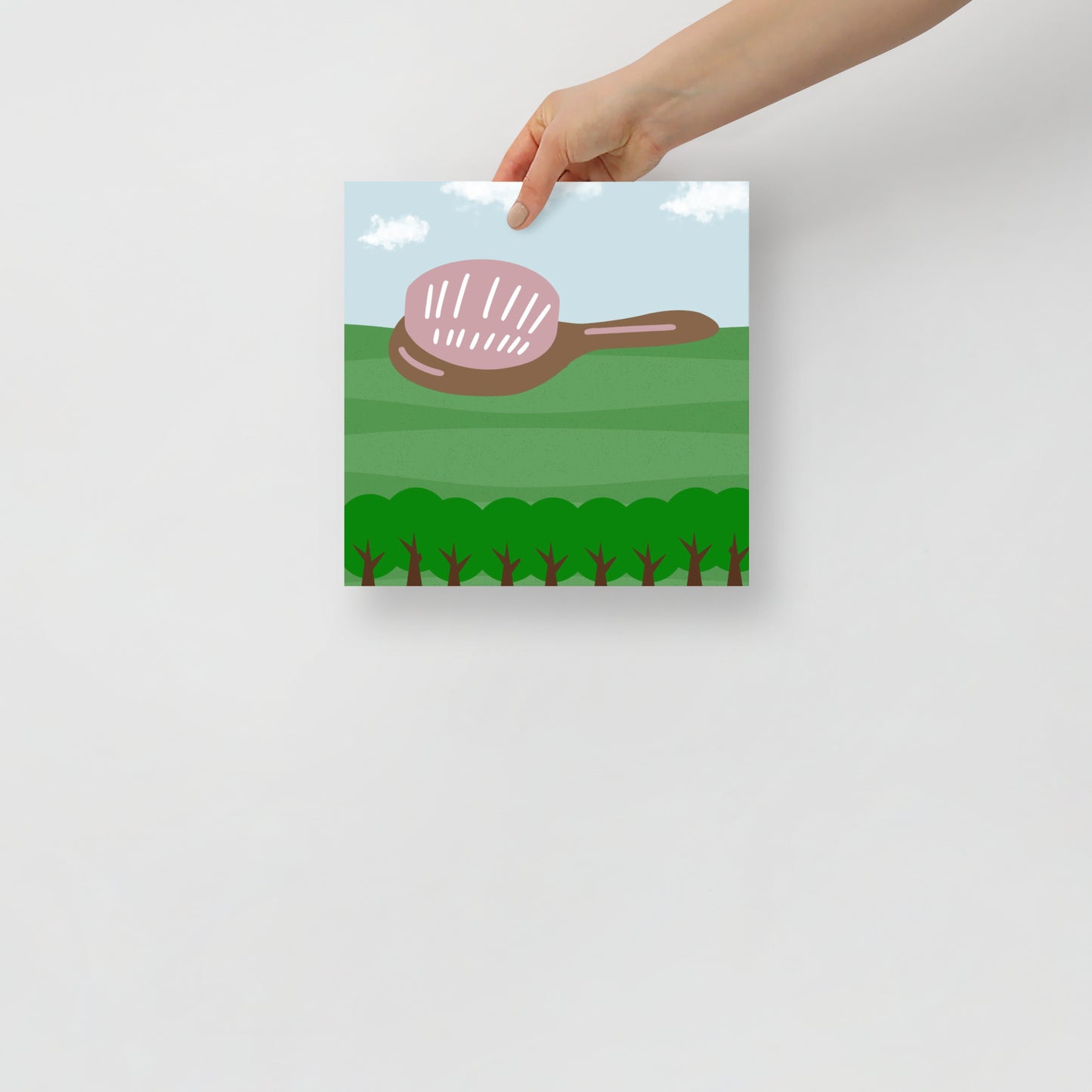 Hand holding a poster with a hair brush illustration on a green field and clouds.