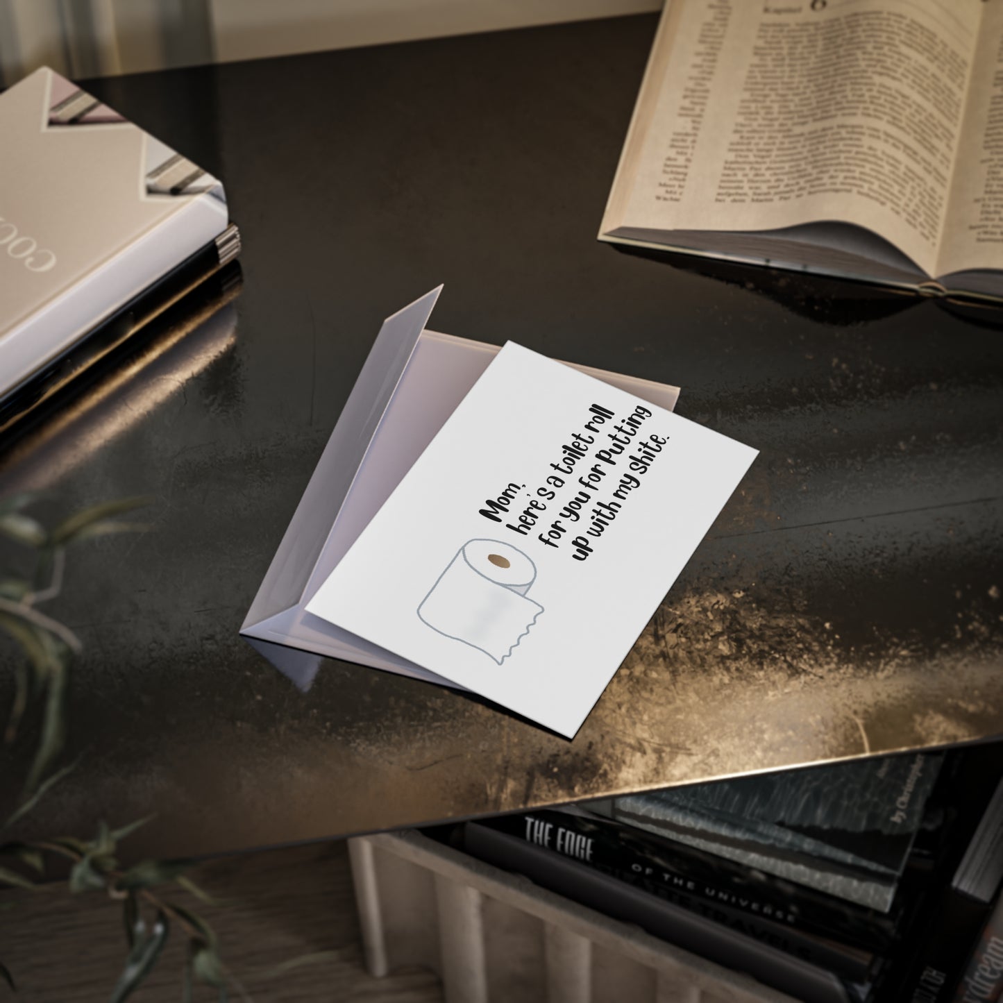 Greeting card with a roll of toilet paper graphic on a dark surface with books and a plant.