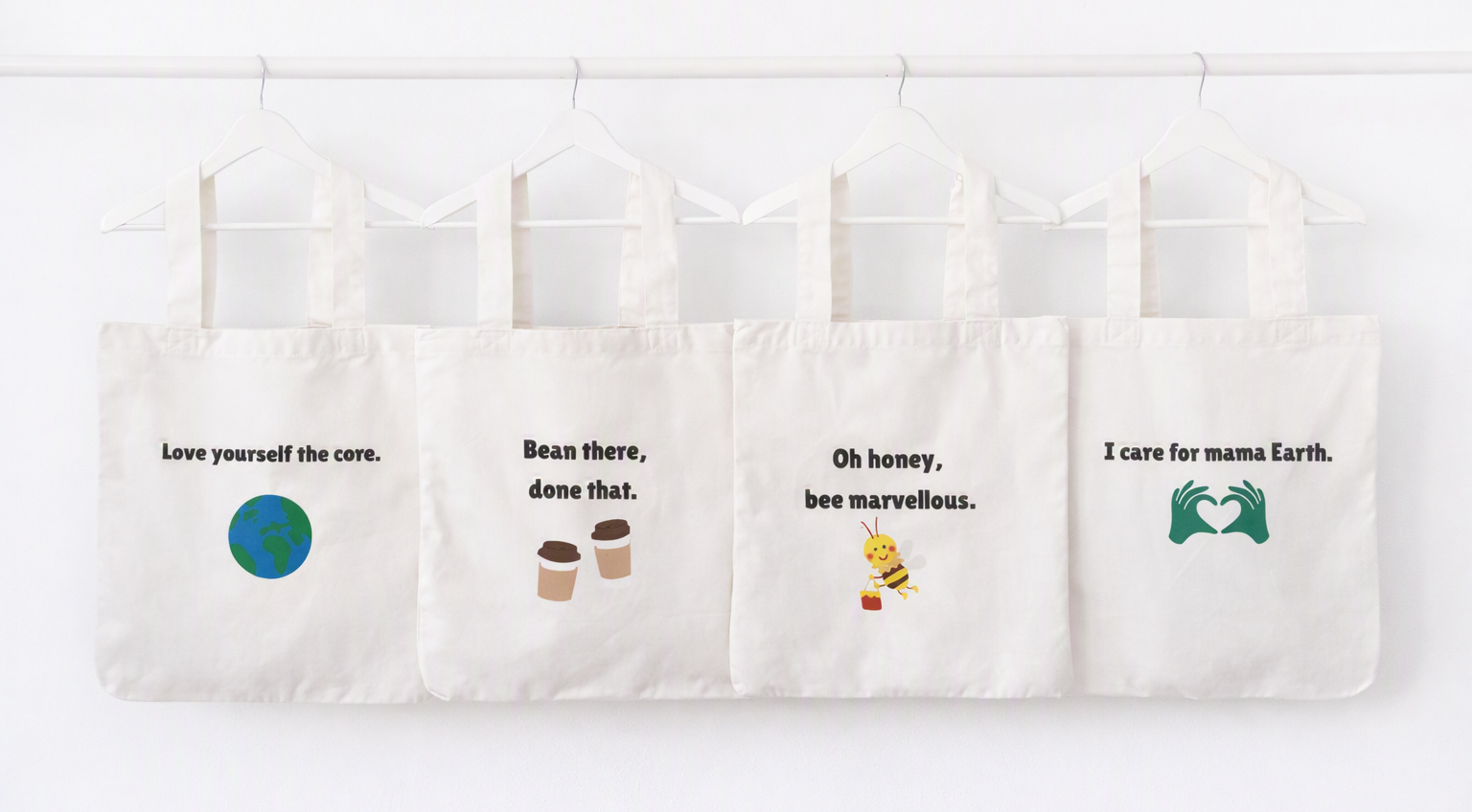 Four white canvas tote bags hanging from a white rod against a white wall. Each bag has a different printed quote and small illustration: 'Love yourself the core.