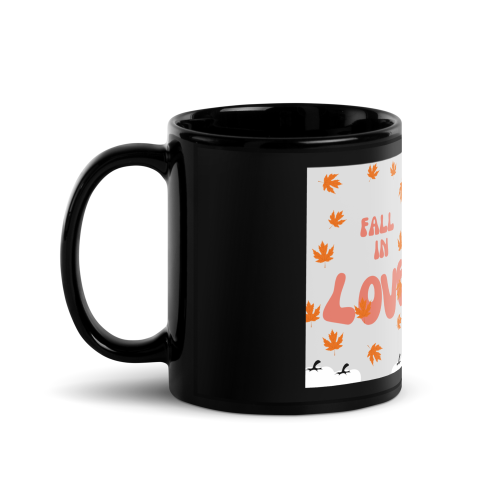 A glossy black ceramic mug with a white printed graphic on the side featuring orange autumn leaves and the text 'FALL IN LOVE'.