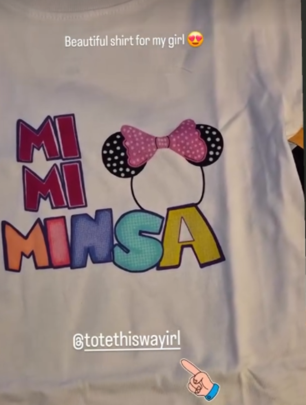 A white short-sleeve t-shirt featuring a colorful custom graphic that reads 'MI MINSA' in multicolored block letters, styled with a Minnie Mouse-inspired head silhouette and a pink polka-dot bow. The photo is taken in a casual setting with social media text overlays.