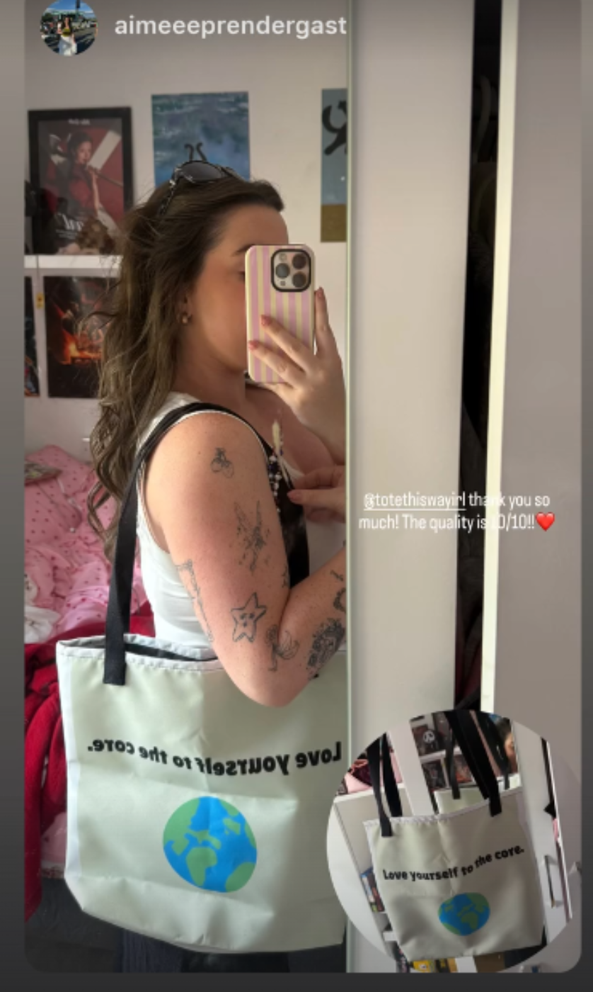 A woman takes a mirror selfie in a bedroom while wearing a white canvas tote bag with black handles and a blue and green globe graphic. The bag has the text 'LOVE YOURSELF TO THE CORE.