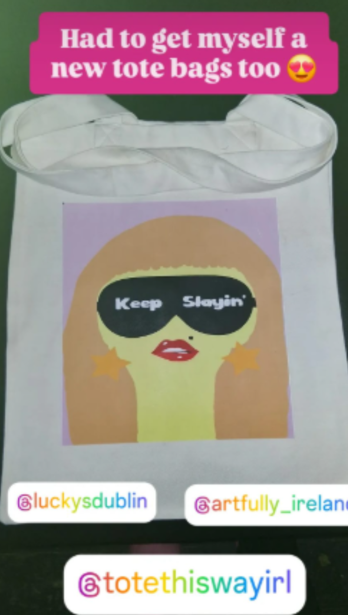 A white tote bag with a colorful graphic of a cartoon figure wearing black sunglasses that say 'Keep Stayin''. The bag is shown against a dark green background with social media-style text and handles overlaid on the image.