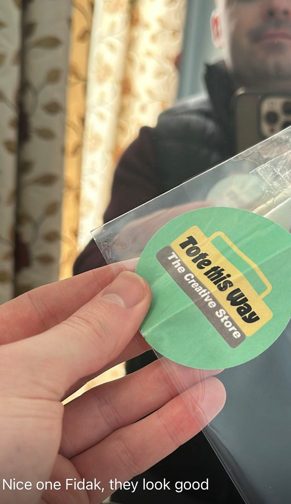 A hand holds a small round sticker inside a clear plastic sleeve. The sticker is light green with a yellow and brown logo that reads 'Tote this Way The Creative Store.