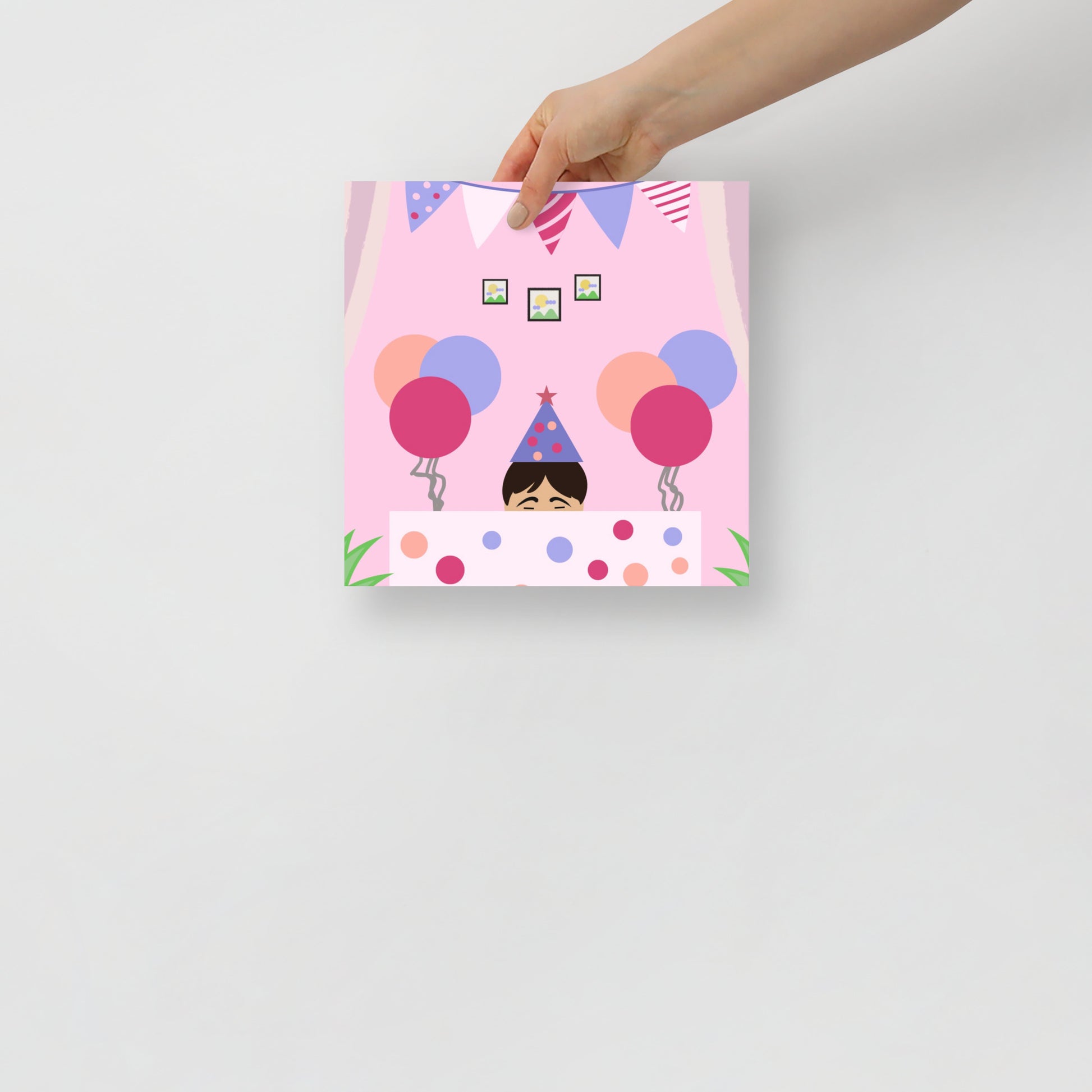 Hand holding a colourful art poster of a child's birthday with balloons, bunting and a party hat on a pink background.