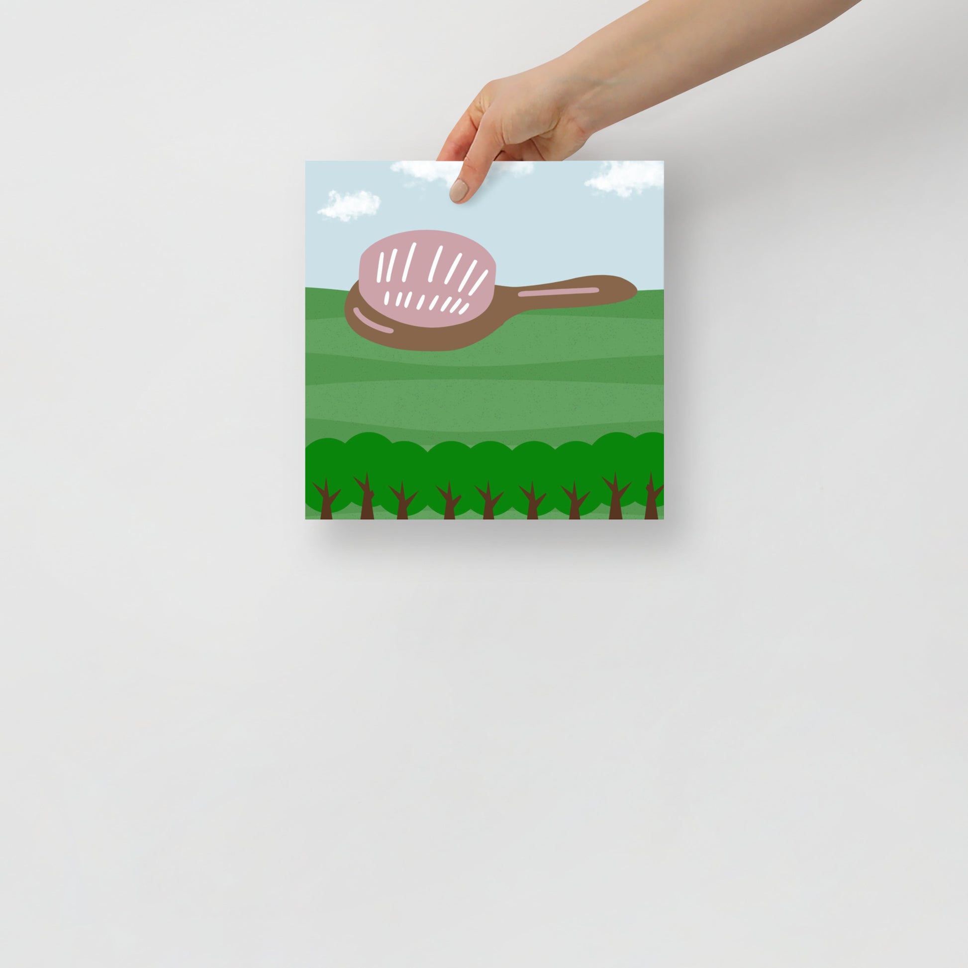 Hand holding a poster with a hair brush illustration on a green field and clouds.