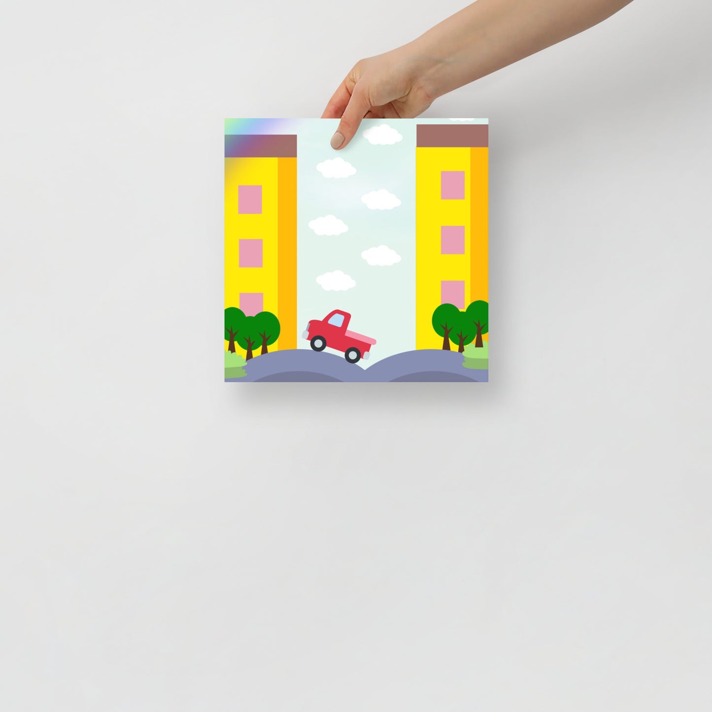Hand holding a small art poster of a cityscape with buildings, trees, and a car on a white background