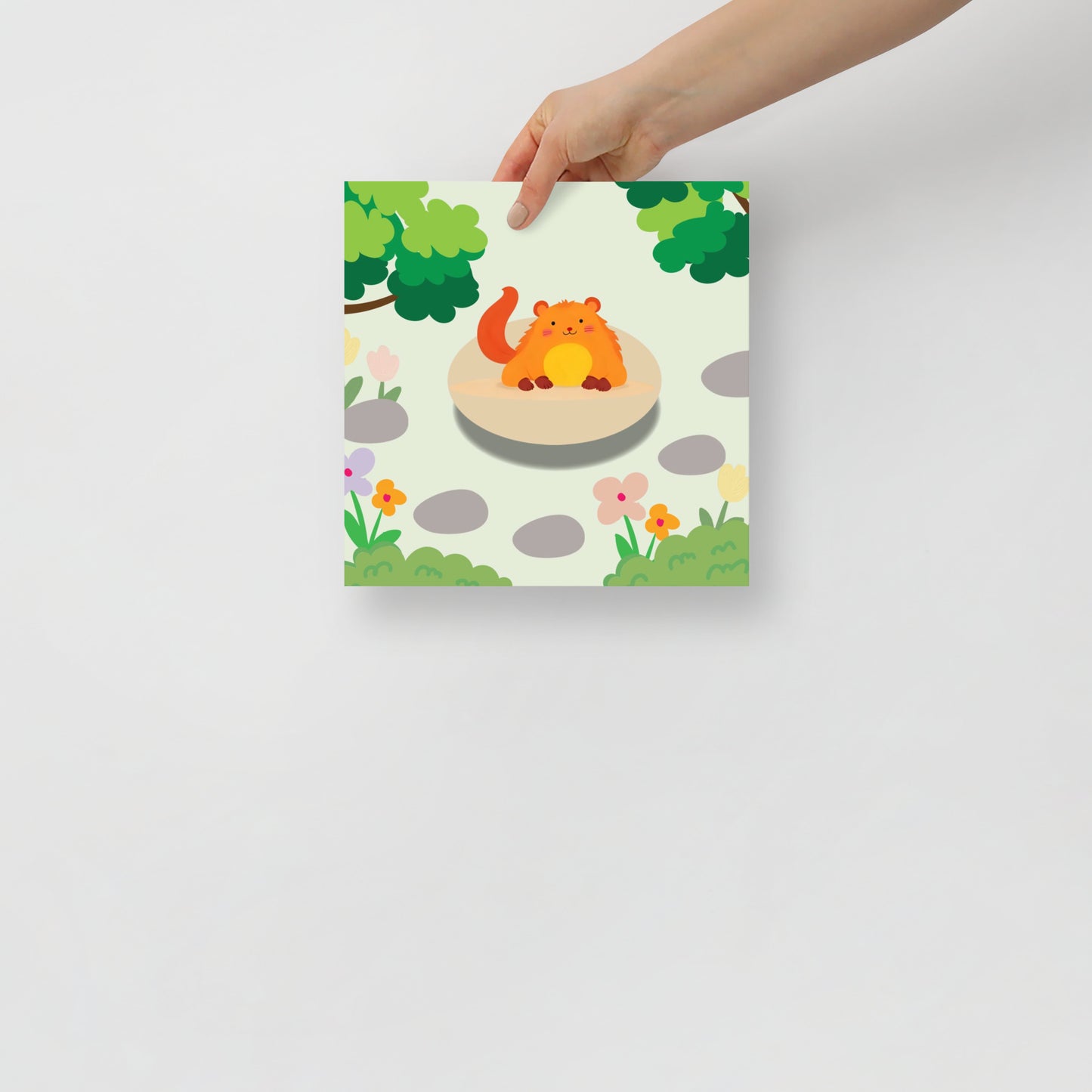 Hand holding a colorful poster with a cartoon critter on a stone in a garden setting.