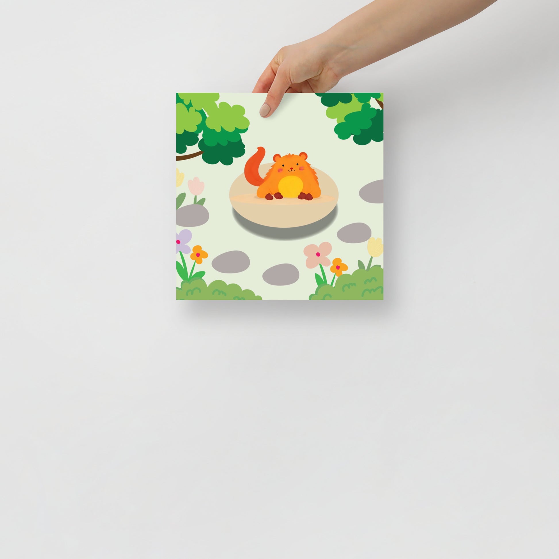 Hand holding a colorful poster with a cartoon critter on a stone in a garden setting.